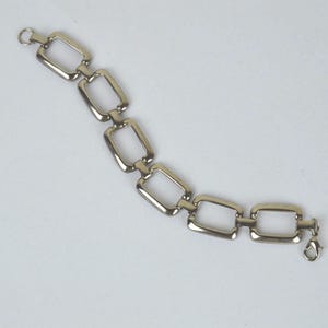 May include: A silver-coloured bracelet with a series of rectangular links. The bracelet has a lobster clasp and a small loop at the other end. The links are connected in a chain-like pattern, creating a modern accessory.