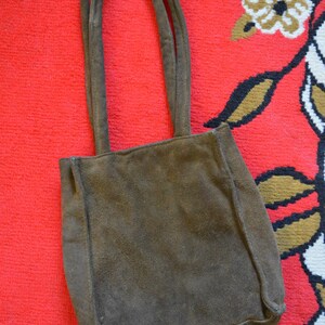 May include: A brown suede tote bag with two handles. The bag is rectangular with a square base and is set against a red patterned background. The bag's color is a muted brown.