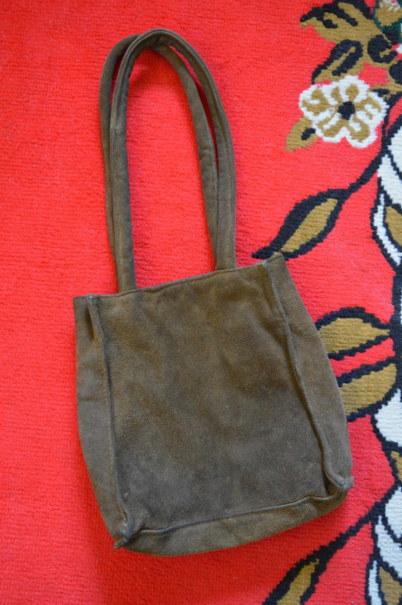 May include: A brown suede tote bag with two handles. The bag is rectangular with a square base and is set against a red patterned background. The bag's color is a muted brown.