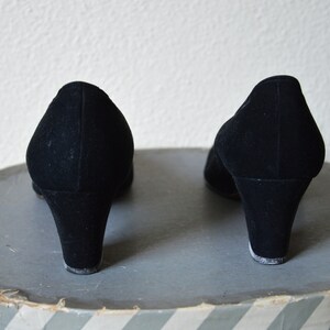 May include: A pair of black suede heeled shoes with a low heel. The shoes are on a gray and white striped surface.