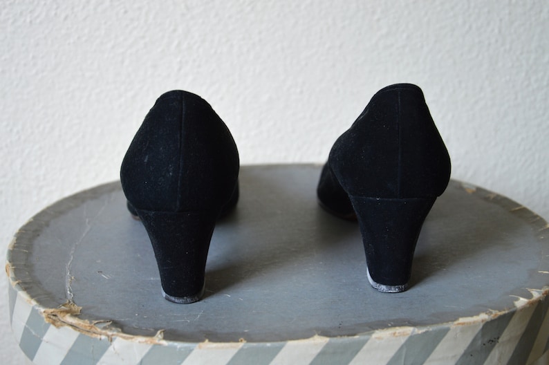 May include: A pair of black suede heeled shoes with a low heel. The shoes are on a gray and white striped surface.