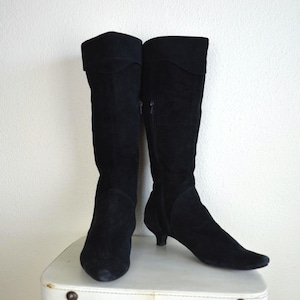 Może przedstawiać: Para czarnych zamszowych kozaków do kolan z niskim obcasem. Buty mają wywinięty mankiet u góry i boczny zamek błyskawiczny. Buty są prezentowane na białej powierzchni na białym tle.