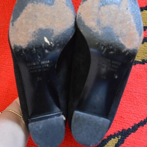 May include: A pair of black suede heels with worn soles. The heels have a block heel design and are size 39. The soles show signs of wear and tear. The shoes are made of leather and are labeled "Made in Italy."