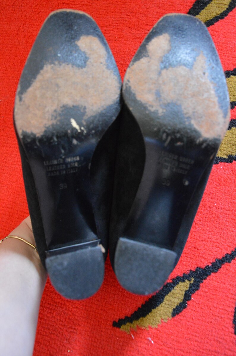 May include: A pair of black suede heels with worn soles. The heels have a block heel design and are size 39. The soles show signs of wear and tear. The shoes are made of leather and are labeled "Made in Italy."