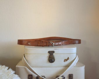 May include: A brown leather belt with a silver buckle rests atop a stack of vintage white suitcases. The belt is a medium brown color and appears to be made of textured leather. The suitcases have a cream-colored finish and a metal clasp.