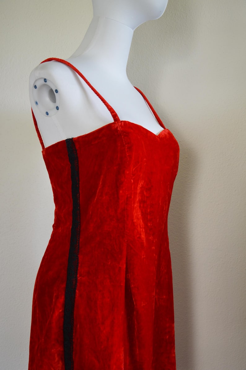 May include: A striking red velvet dress featuring delicate spaghetti straps and a sweetheart neckline. A black stripe adorns the side. The dress is presented on a white mannequin.