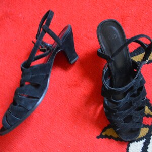 May include: A pair of black suede sandals with open toes and a strap around the ankle. The sandals have a small heel and are displayed on a red surface.
