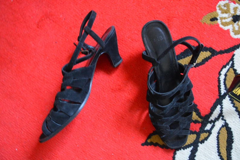 May include: A pair of black suede sandals with open toes and a strap around the ankle. The sandals have a small heel and are displayed on a red surface.