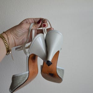 Puede incluir: Un par de zapatos de tac&oacute;n destalonados en gris claro con un patr&oacute;n texturizado similar a la piel de serpiente en la puntera, tacones de cuero liso y finas correas ajustables en el tobillo. Las suelas son color canela con desgaste.