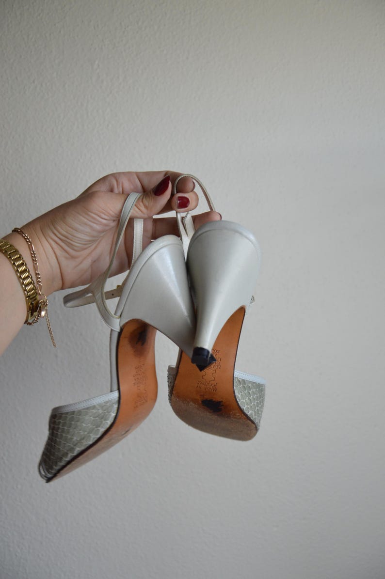 Puede incluir: Un par de zapatos de tac&oacute;n destalonados en gris claro con un patr&oacute;n texturizado similar a la piel de serpiente en la puntera, tacones de cuero liso y finas correas ajustables en el tobillo. Las suelas son color canela con desgaste.