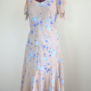 May include: A light peach-coloured dress with a floral print featuring blue and light blue roses. The dress has short sleeves with a tie detail at the shoulders and a flowing skirt.