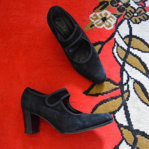May include: A pair of black suede Mary Jane style shoes with a block heel. One shoe is positioned above the other on a red surface. The shoes have a strap across the top of the foot and a rounded toe.