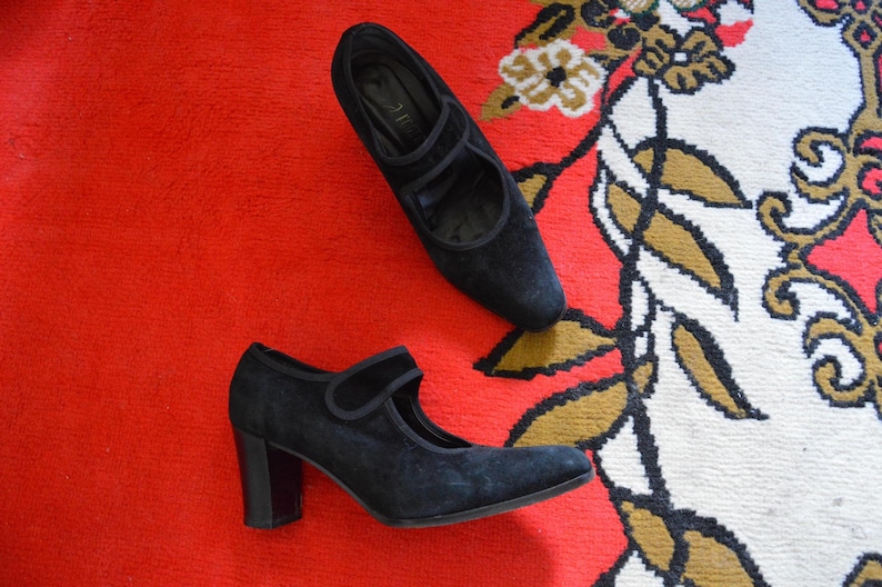 May include: A pair of black suede Mary Jane style shoes with a block heel. One shoe is positioned above the other on a red surface. The shoes have a strap across the top of the foot and a rounded toe.