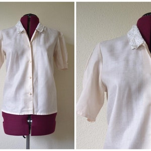 Puede incluir: Una camisa blanca de botones con cuello de encaje. La camisa tiene mangas cortas y un corte holgado.