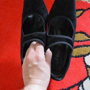 May include: A pair of black suede shoes with a pointed toe and a strap across the top. The shoes are on a red patterned rug. The shoes have a heel and a cutout design.
