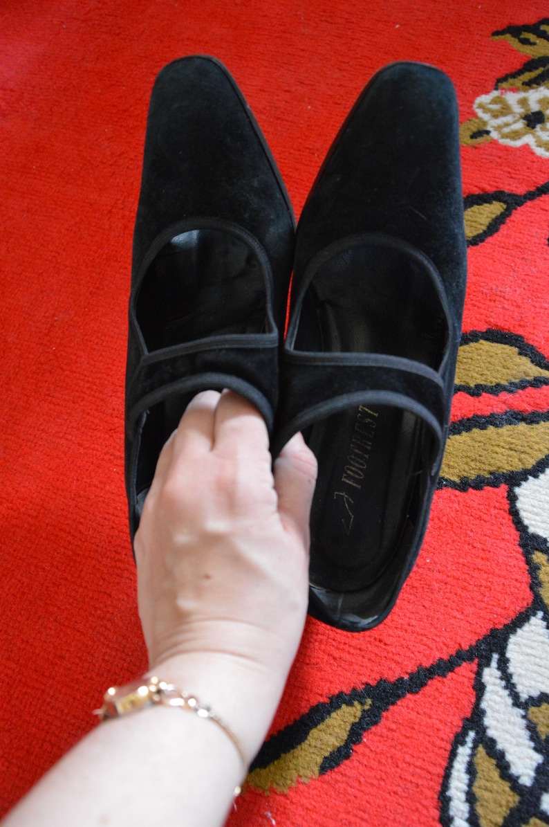 May include: A pair of black suede shoes with a pointed toe and a strap across the top. The shoes are on a red patterned rug. The shoes have a heel and a cutout design.