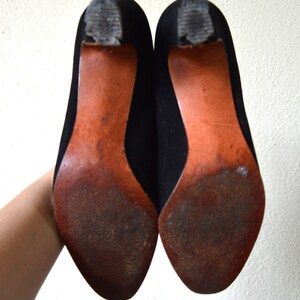 May include: A pair of black suede heeled shoes with brown leather soles. The shoes are worn and have some scuffs on the soles.
