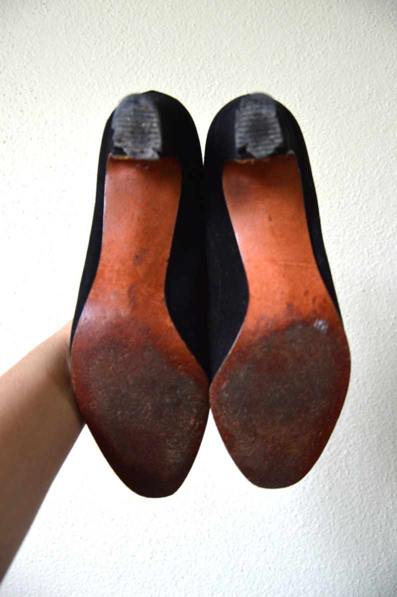 May include: A pair of black suede heeled shoes with brown leather soles. The shoes are worn and have some scuffs on the soles.