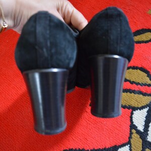 May include: Pair of black suede shoes with a rounded toe and a block heel. The heels are approximately 3 inches tall. The shoes are held against a red patterned background.