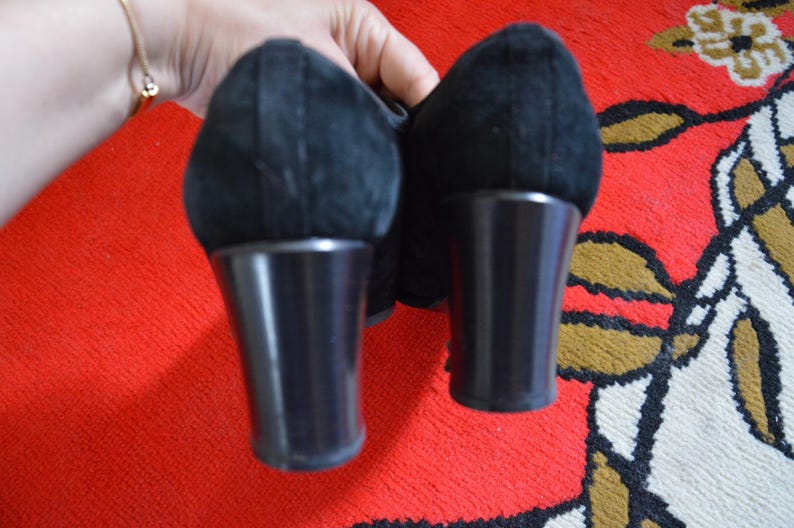 May include: Pair of black suede shoes with a rounded toe and a block heel. The heels are approximately 3 inches tall. The shoes are held against a red patterned background.
