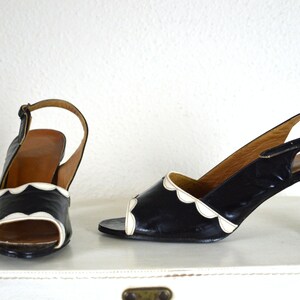 May include: A pair of black heeled shoes with white scalloped trim around the toe and ankle strap. The shoes are on a white surface.