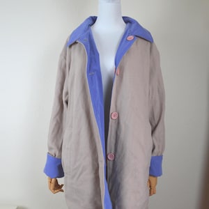 Könnte beinhalten: Ein hellbrauner Mantel mit blauem Kragen, Manschetten und Innenbesatz. Der Mantel hat rosa Knöpfe und wird auf einer Schaufensterpuppe präsentiert. Der Mantel ist ein langer, lässiger Stil.