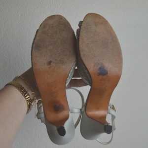Puede incluir: Un par de sandalias de tac&oacute;n alto de color claro con correas al tobillo, vistas desde abajo. Las suelas son de color tostado con desgaste visible, acabado texturizado, tac&oacute;n fino y texto estampado que indica el cuero y origen.