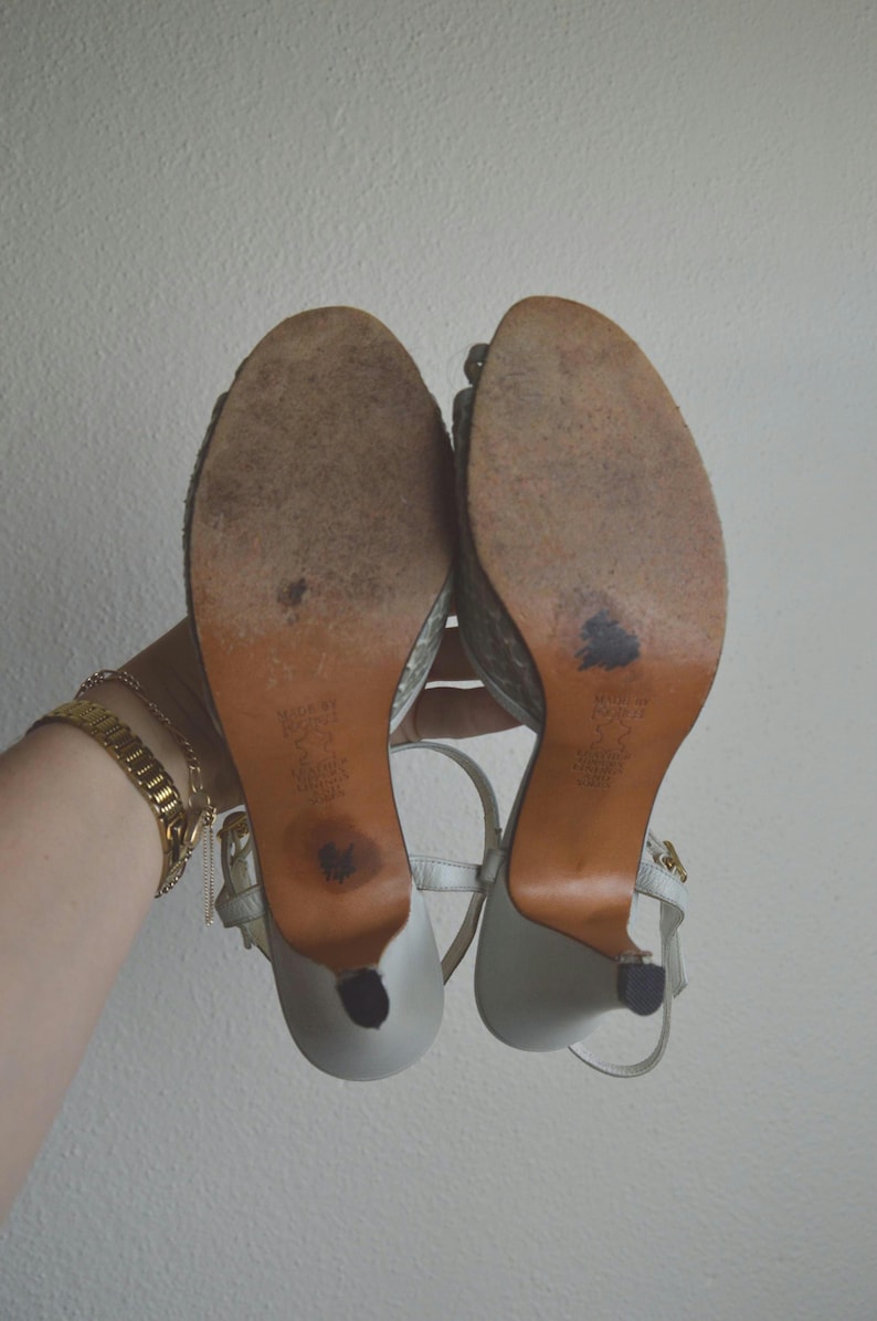 Puede incluir: Un par de sandalias de tac&oacute;n alto de color claro con correas al tobillo, vistas desde abajo. Las suelas son de color tostado con desgaste visible, acabado texturizado, tac&oacute;n fino y texto estampado que indica el cuero y origen.