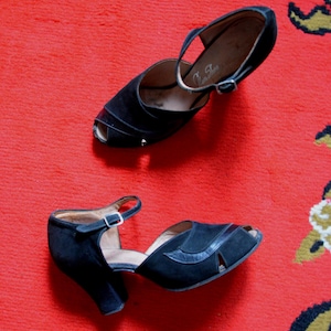 Peut inclure: Une paire d'escarpins noirs vintage à bout ouvert avec une bride et une fermeture à boucle. Les chaussures ont un talon incurvé et sont posées sur un fond rouge à motifs. La marque "Town Shoes" est visible sur la semelle intérieure.