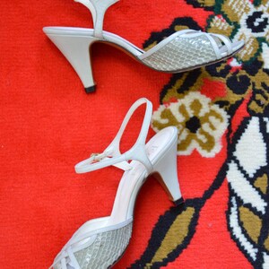 Puede incluir: Un par de zapatos de tac&oacute;n destalonados blancos con un panel frontal texturizado con patr&oacute;n de piel de serpiente gris claro, dise&ntilde;o de punta abierta, una correa de tobillo delgada con una peque&ntilde;a hebilla dorada y tac&oacute;n medio con tapa negra.