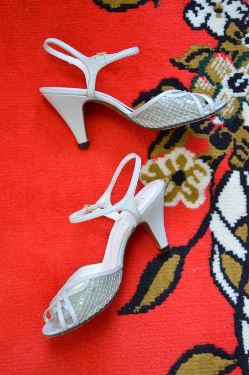 Puede incluir: Un par de zapatos de tac&oacute;n destalonados blancos con un panel frontal texturizado con patr&oacute;n de piel de serpiente gris claro, dise&ntilde;o de punta abierta, una correa de tobillo delgada con una peque&ntilde;a hebilla dorada y tac&oacute;n medio con tapa negra.