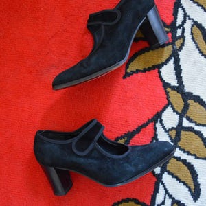 May include: A pair of black suede Mary Jane style shoes with a block heel. The shoes have a strap across the top of the foot and a red sole. The shoes are on a red and patterned surface.