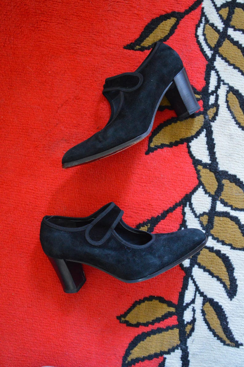 May include: A pair of black suede Mary Jane style shoes with a block heel. The shoes have a strap across the top of the foot and a red sole. The shoes are on a red and patterned surface.
