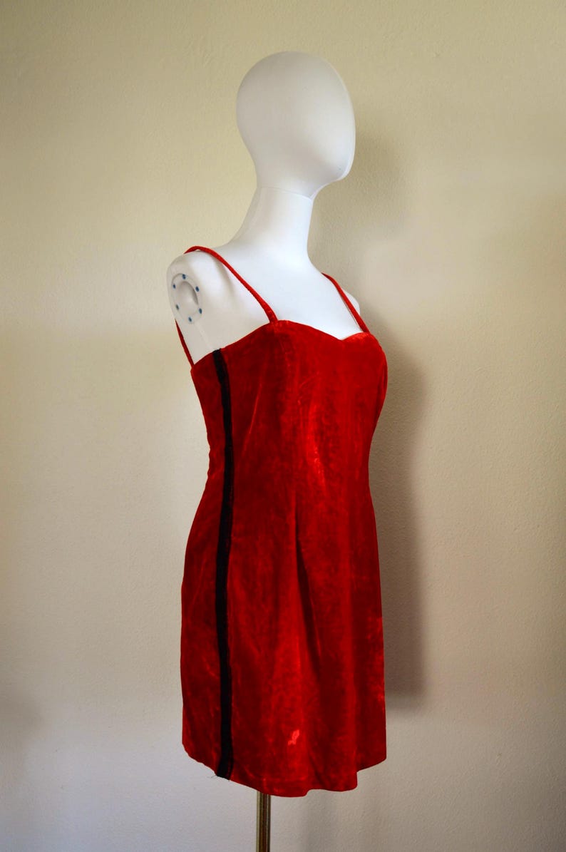May include: A red velvet dress with thin straps and a sweetheart neckline. A black vertical stripe runs down the left side of the dress. The dress is displayed on a white mannequin.