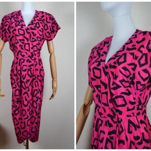 May include: A vintage wrap dress in a vibrant pink hue, adorned with a bold black geometric pattern. The dress features short sleeves, a V-neckline, and a fitted waist, creating a flattering silhouette. The dress is displayed on a mannequin.