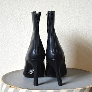 May include: A pair of black leather ankle boots with a zipper on the back of each boot. The boots have a high heel and are sitting on a striped surface.