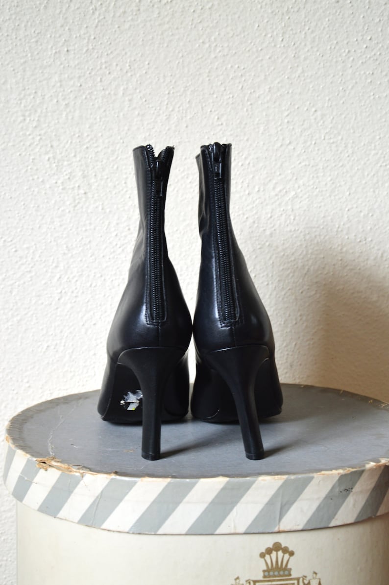 May include: A pair of black leather ankle boots with a zipper on the back of each boot. The boots have a high heel and are sitting on a striped surface.