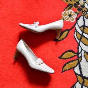 Puede incluir: Un par de zapatos de salón de cuero blanco con punta afilada y tacón pequeño. Los tacones tienen aspecto de madera. Los zapatos están sobre una alfombra roja con un estampado floral en dorado, negro y blanco.