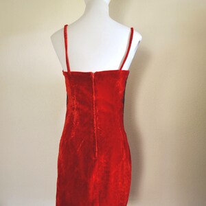 May include: A red velvet dress with thin spaghetti straps and a straight neckline. The dress is displayed on a white mannequin, showcasing its form-fitting silhouette and the vertical zip detail down the back.