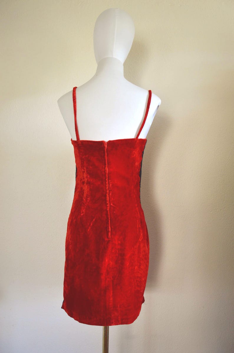 May include: A red velvet dress with thin spaghetti straps and a straight neckline. The dress is displayed on a white mannequin, showcasing its form-fitting silhouette and the vertical zip detail down the back.