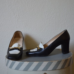 Könnte beinhalten: Ein Paar schwarze und weiße Schuhe mit Absatz und goldenem Schnallen-Detail. Die Schuhe stehen auf einer gestreiften Schachtel.