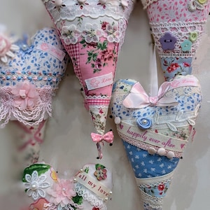 May include: Five handmade fabric heart decorations with lace, ribbons, and buttons. The hearts are in shades of pink, blue, and white. The text on the hearts reads "Love", "Happily Ever After", and "All My Love".