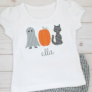 May include: A white short-sleeved t-shirt featuring a ghost, an orange pumpkin, and a black cat in gingham patterns, with the name "ella" embroidered in black script below the designs, paired with black and white gingham ruffled bloomers.