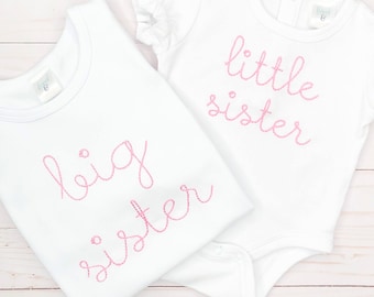 May include: Two white baby shirts with pink embroidered text. One shirt says "big sister" and the other says "little sister". The "little sister" shirt has ruffled sleeves. Both shirts are on a light wood surface.