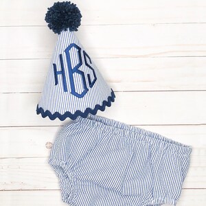 Pode incluir: Um conjunto de beb&eacute; azul e branco listrado com um chap&eacute;u azul com pompom. O chap&eacute;u tem um monograma azul que diz "HBS".
