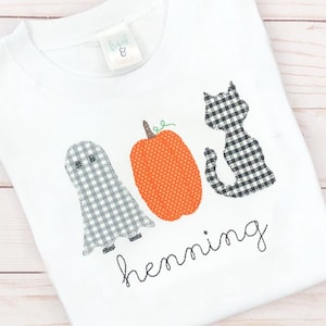 May include: A white t-shirt featuring an embroidered design of a grey gingham ghost, an orange textured pumpkin, and a black and white gingham cat. The name "henning" is embroidered in black cursive script below the figures. A tag reads "frank & design".