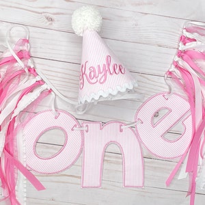 Puede incluir: Una pancarta a rayas rosa y blanca con la palabra "one" en letras grandes de color rosa. La pancarta está decorada con cintas rosa y blancas. Se incluye un gorro de fiesta a rayas rosa y blancas con el nombre "Kaylee" bordado.