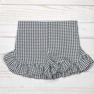 May include: A pair of shorts featuring a black and white gingham check pattern. The garment has a high waistband and a wide, gathered ruffle trim along the bottom hemline that extends across the front and sides of the leg openings.