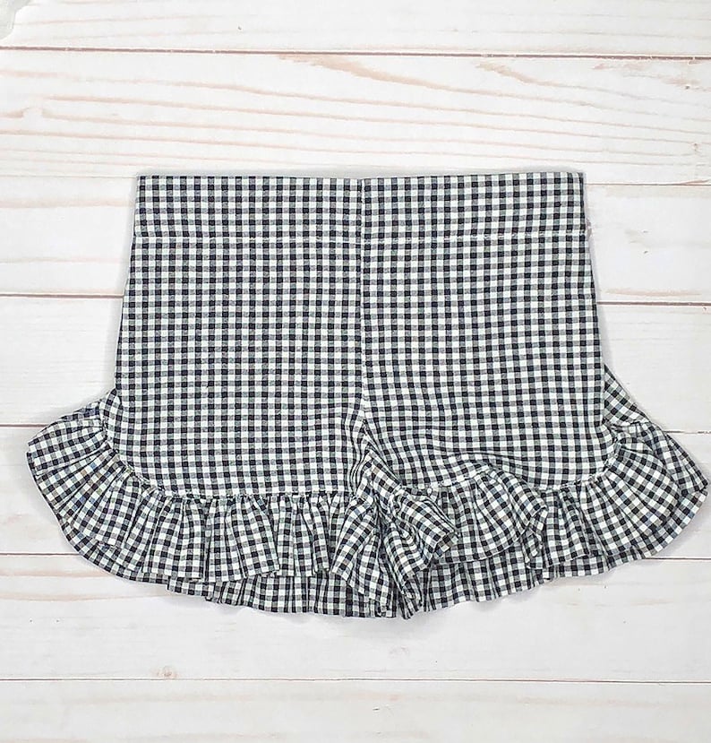 May include: A pair of shorts featuring a black and white gingham check pattern. The garment has a high waistband and a wide, gathered ruffle trim along the bottom hemline that extends across the front and sides of the leg openings.