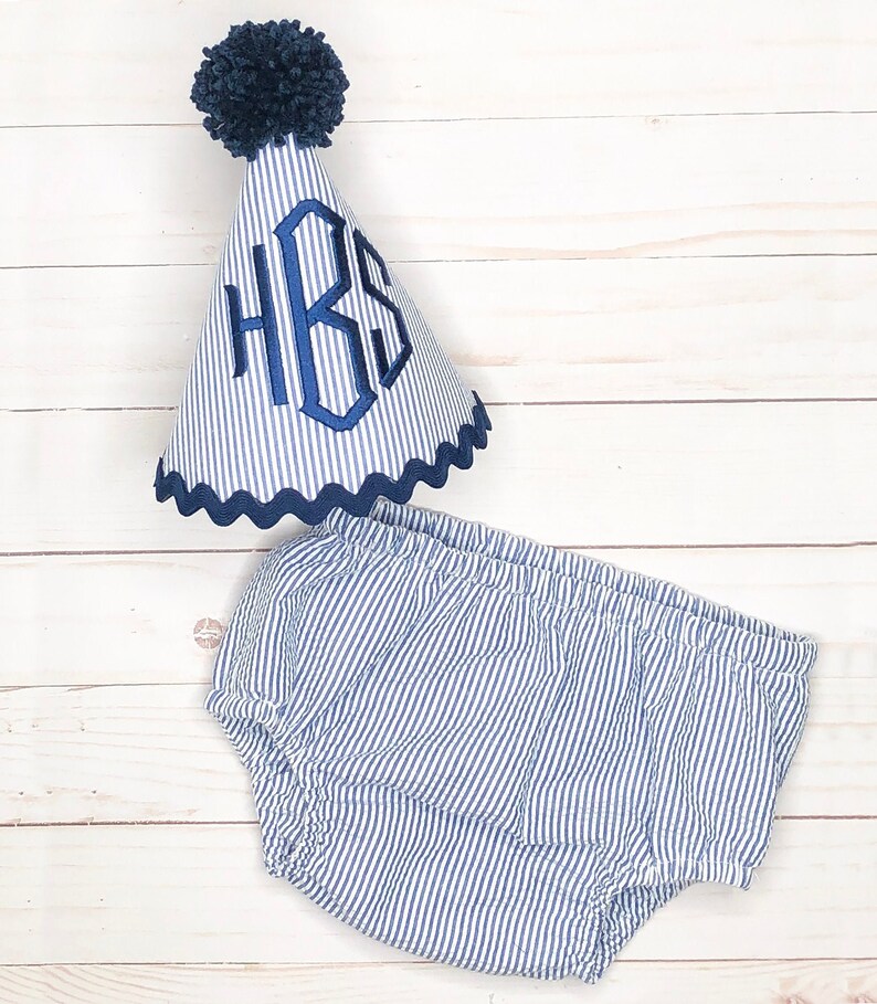 Pode incluir: Um conjunto de beb&eacute; azul e branco listrado com um chap&eacute;u azul com pompom. O chap&eacute;u tem um monograma azul que diz "HBS".